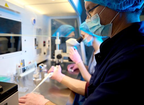 Two embryologists in protective clothing and masks work together in a sterile lab, one holding a vial